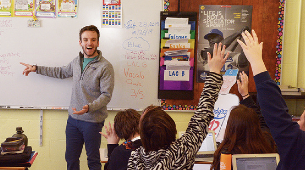 Teacher in a classroom pointing a whiteboard with students sitting at desks with their hands raised.